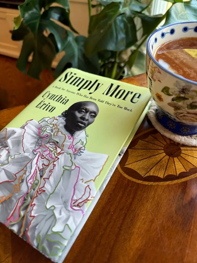 wood tabletop with a book titled "Simply More" by Cynthia Erivo and a flowered coffee cup to the right. A green plant stands behind the table.