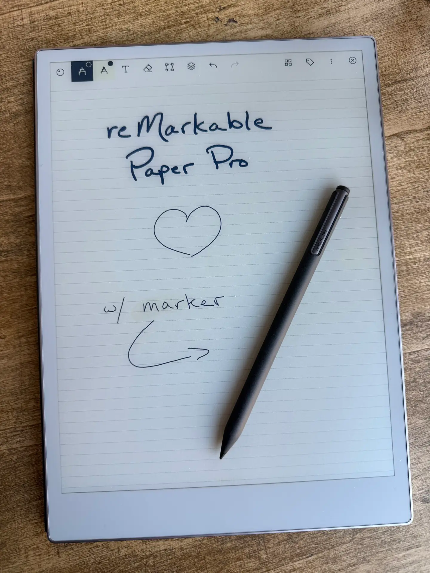 a picture of a reMarkable Paper Pro, with the message "reMarkable Paper Pro" with a sketch of a heart below it, with "w/ marker" and a drawn arrow pointing to the stylus resting on the screen. The tablet is resting on a wooden surface.