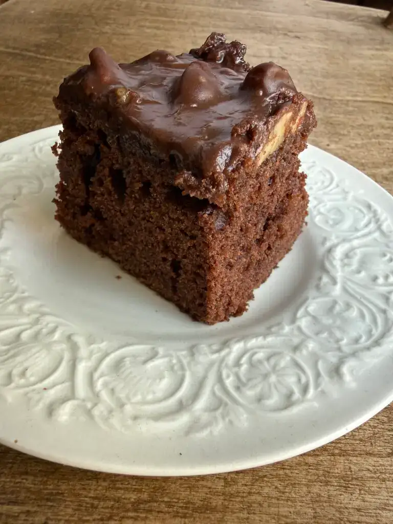 Chocolate Sheet Cake - a slice sits on top of a cream plate with a textured scroll detail around the rim. The plate sits on a wood table top.