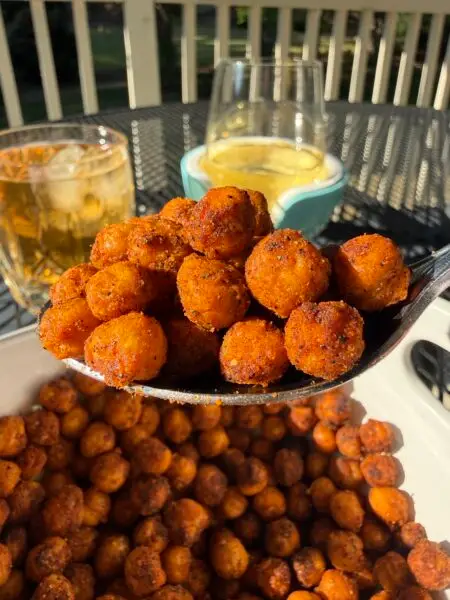 Set on a black wrought iron garden table on a sunny day is a bowl of golden, crispy roasted chickpeas. A spoonful of chickpeas is being raised above the bowl, and two cocktails are set on the table behind the bowl.