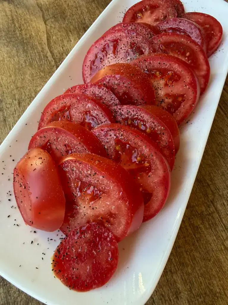 Fresh sliced red tomatoes, sprenkled with sea salt and ground pepper, arranged on a white rectangle platter, which sits on a wood table.