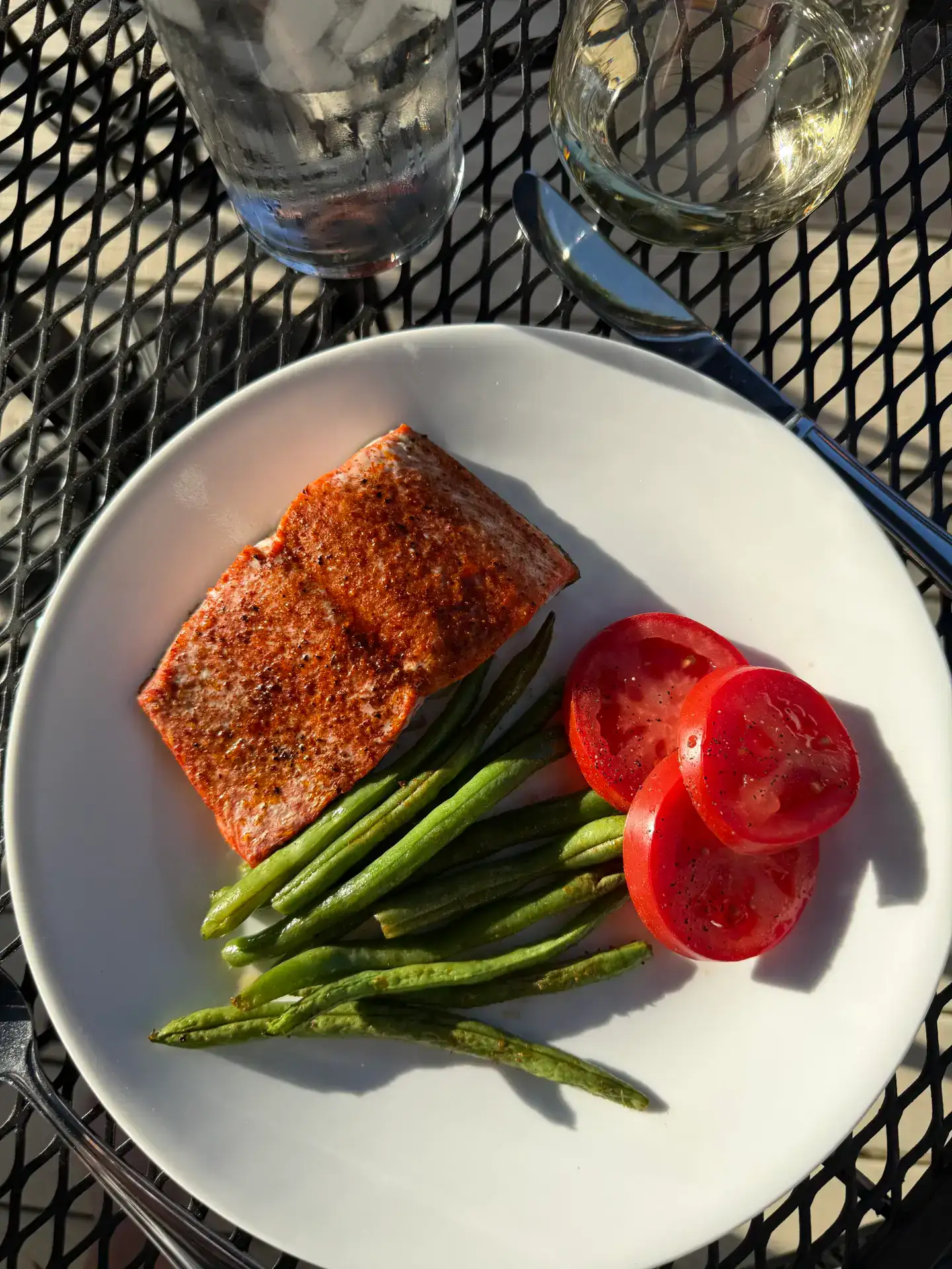 Roasted Salmon and Green Beans on a white plate with red sliced tomatoes, sitting on a black iron tabletop, with a glass of water and a glass of wine visible at the top of the picture. The picture is taken outside on a sunny day.