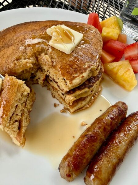a stack of whole wheat pancakes with a bite cut out, with melting butter and syrup on top, alongside fruit salad and sausage links on a white plate.