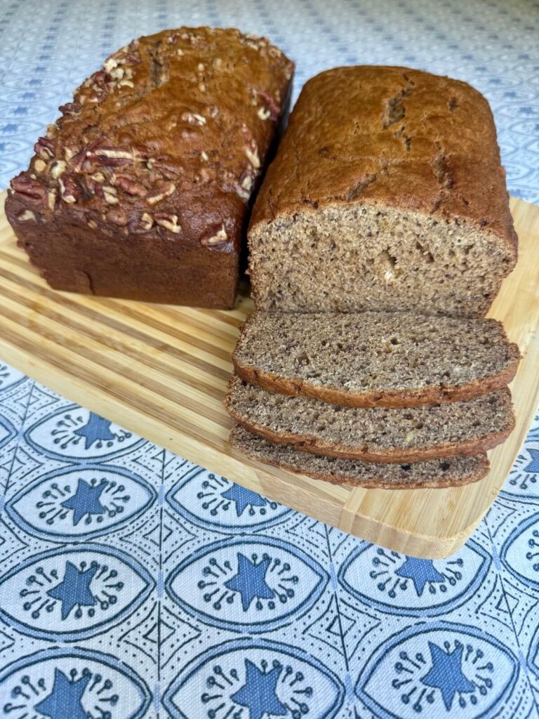 Two finished loaves of Mom's Whole Wheat Banana Bread on a wooden cutting board, showing a pecan-topped loaf alongside a plain loaf sliced to reveal its moist interior.
