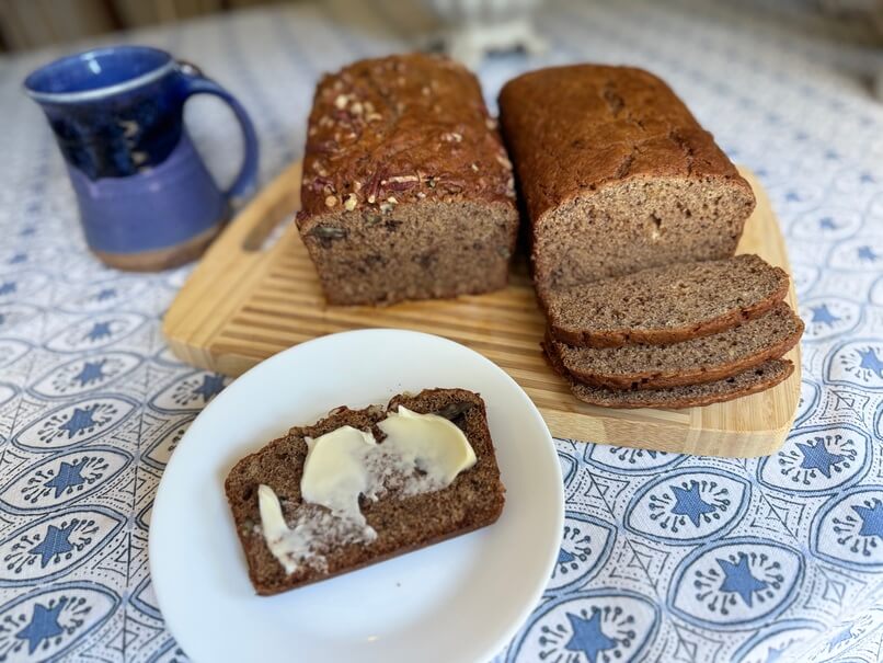 Two loaves of Mom's Whole Wheat Banana Bread, one with pecans and one without, served on a wooden cutting board with a buttered slice on a white plate.