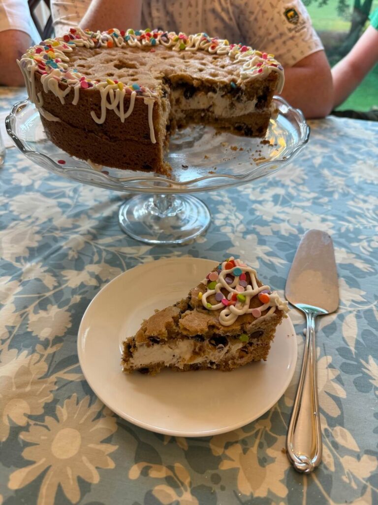 Layered chocolate chip cookie cake on a glass cake stand with a slice cut out. The slice of cookie cake is on a white plate in front, with a cake server to the right.